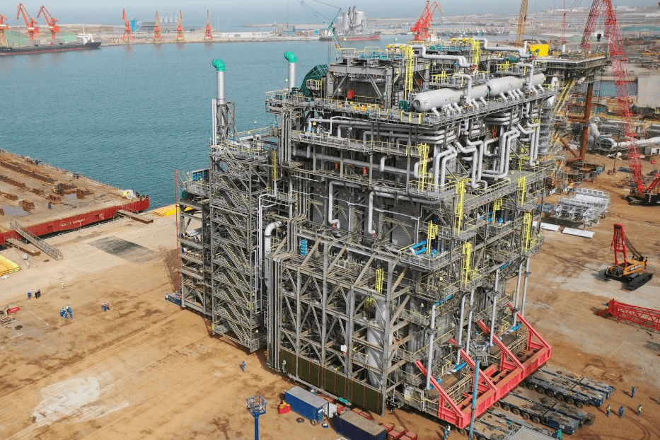 Large steel industrial structure under construction at a waterfront site, with scaffolding, cranes and construction materials surrounding the work area.